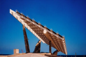 A large, modern solar panel structure with an angular design, supported by several inclined columns, stands against a clear blue sky. The geometric pattern of the pérgola fotovoltaica creates strong lines and shadows, emphasizing the architectural form.