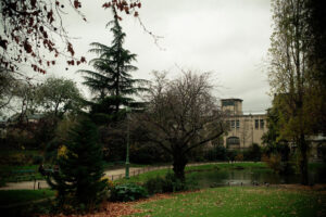 A tranquil scene at Square des Batignolles on a cloudy day features a small pond surrounded by trees, some with fallen autumn leaves. A pathway winds through the park, with a few benches and ducks near the water. A large building stands in the background.