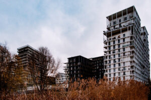 A cluster of modern, multi-story apartment buildings stands under a cloudy sky in Parc Clichy-Batignolles. The buildings have balconies and an architectural design featuring white and dark-colored exteriors. Bare trees and tall reeds are in the foreground, suggesting a late autumn or early winter setting.