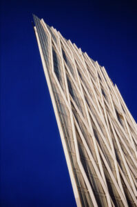 A tall, modern skyscraper with a unique crisscross pattern on its facade rises sharply against a deep blue sky in Barcelone. The Diagonal Zero Zero building's sleek, angular design is accentuated by the sunlight, casting shadows that highlight its architectural details.