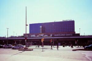 A large building labeled "Barceló Sants" rises above Barcelona Sants railway station. The station's entrance displays "BARCELONA SANTS" in bold letters. Streets and parked cars are visible in front, and a few people are walking nearby in the heart of Barcelona.