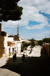 A narrow, sunny street lined with low buildings, trees, and parked motorcycles stretches through the serene Carrer de Marià Labèrnia. A tall communication tower stands in the background against a partly cloudy blue sky near Turó de la Rovira. The street appears calm and quiet in Barcelone.