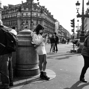Black and white photo of a person wearing a mask, coat, skirt, and boots, standing against a stone structure on the bustling Place de l'Opéra. They are looking at their phone while people walk by. In the background, there are historic buildings and streetlights.
