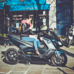 A person sits on a parked blue motorcycle in front of a restaurant, glancing at their smartphone. They are wearing a black jacket with a pink scarf and beige pants. Another scooter is parked nearby, and the restaurant has signs for various services.