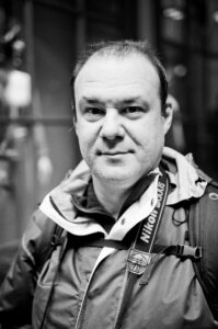 Black and white photo of a middle-aged man wearing a jacket and a backpack. He has a Nikon camera strap around his neck, looking directly at the camera with a neutral expression. The blurred background hints at an urban setting, capturing the essence of street photographer Fabrice Mercier.