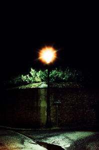 A single streetlamp casts a warm, bright light in the dark night, illuminating adjacent greenery and part of a cobblestone street. A sign on a nearby wall reads "Rue de la Charette" in Nogent-Le-Rotrou. The overall scene exudes a quiet, serene atmosphere, perfectly capturing the essence of nuit.