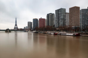 A cityscape featuring the Eiffel Tower in the background on a cloudy day. High-rise buildings of Beaugrenelle line the riverbank on the right, with several docked boats along the water. The river appears calm, reflecting the overcast sky.