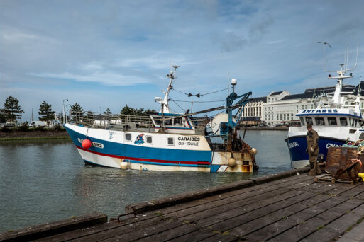 A fishing boat named "Caraibes" is docked at a harbor, with another boat called "Antares" nearby. The scene includes calm water, a cloudy sky, a wooden pier in the foreground, and a few people standing on the pier, possibly working or preparing to board.