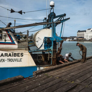 Three individuals work on maintenance at the edge of a wooden pier next to a blue and white fishing boat named "Caraïbes". In the background, there is a calm body of water and a large white building. The sky is clear and light blue.