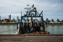 A fishing boat docked at a pier, equipped with large reels and various fishing equipment. The background features buildings, trees, and a slightly overcast sky. The water reflects some of the surrounding scenery.