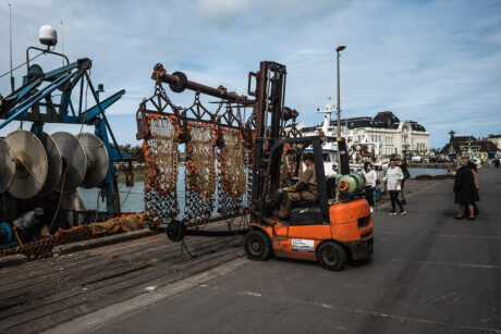 A forklift transports a load of fishing nets and equipment along a dock. People walk nearby, and various maritime buildings and vessels are visible in the background under a partly cloudy sky.