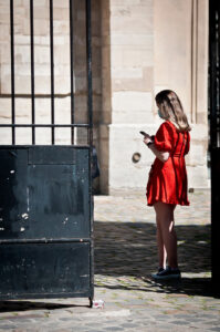 A woman in a robe rouge, wearing a mask, stands on a cobblestone path near a large black iron gate. She is looking at her phone. The background features an old stone building with sunlight casting shadows.
