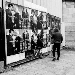 A person in a hoodie stands in front of a wall covered with black-and-white affiches advertising "The Kooples." The posters, collés meticulously, feature various couples in stylish poses against urban backgrounds. The scene is rendered in black and white, suggesting a gritty, urban setting.