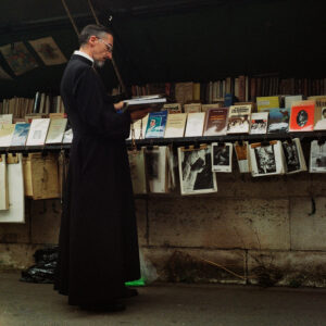 A man in a black long robe, possibly Le père François, stands on a sidewalk in front of an outdoor bookstall, browsing through books. The bookstall displays an array of books and prints hanging on a wire above them. The background is shaded and slightly dim.