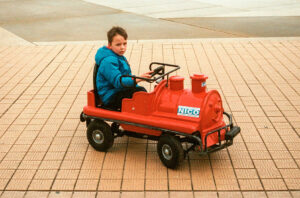 A child wearing a blue jacket is sitting in a red, train-shaped ride-on toy with "NICO" written on it. The scene appears to be outdoors on a paved surface, reminiscent of the playful days Charles Joseph cherished.