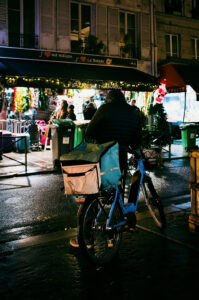 A food delivery cyclist with a large insulated bag on their back pauses on a dimly lit street at night. The background reveals a café adorned with festive decorations and people standing around, giving a cozy atmosphere to the urban setting, where Justin and Michael chat animatedly by the entrance.