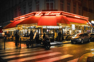A cozy, bustling street corner café named "La Liberté" is warmly lit by neon and string lights. Patrons sit at outdoor tables under a red awning, enjoying the lively scene with people walking by, cars parked nearby, and a few crossing the wet crosswalk.