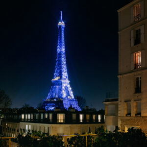 A night view of the Eiffel Tower in Paris illuminated in blue lights, with neighboring buildings softly lit. The sky is clear, adding a serene backdrop to the iconic landmark. Trees and shrubbery can be seen in the foreground, creating a picturesque setting for an unforgettable night tour.