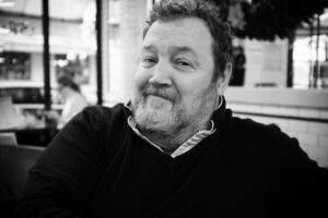 Black and white image of Marc Lepage, a bearded man smiling softly while sitting indoors. The background shows a blurred interior of what appears to be a cafe or restaurant, with another person seated in the distance. Marc wears a sweater over a collared shirt.