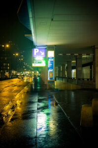 Nighttime street scene with illuminated parking signs under a shelter on the right. Wet pavement reflects the streetlights, and buildings with lit windows are visible in the background. A few cars travel down the road, creating a moody nuit urbaine atmosphere.