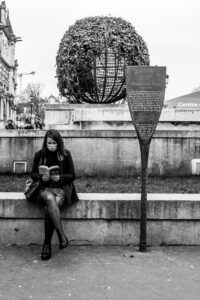 A woman wearing a mask sits on a stone bench reading a book in front of a spherical wire sculpture adorned with vegetation. A signpost with text stands to her right. The background includes historic architecture, an old hôtel, and a construction crane. Black and white photo.