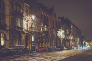 A nighttime urban street scene in Bruxelles featuring historic buildings with illuminated windows. Cars are parked along the curb, and streetlights cast a warm glow on the cobblestone road. The quiet street creates a calm and serene atmosphere, capturing the essence of Belgique's charm.
