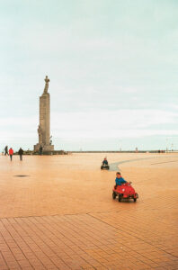 A large plaza in Ostende, Belgique, features a tall statue of a person on a pedestal in the background. Children ride small, red toy cars on the tiled ground. Several people walk in the distance under an overcast sky. The scene captures a calm, open public space with casual activity.