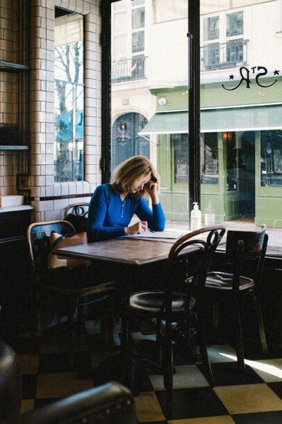 A woman with blonde hair, wearing a blue sweater, sits alone at a wooden table in a dimly lit café beside a large window. She is writing or drawing in a notebook, resting her head on one hand. The café has white tiled walls, and the view outside shows green storefronts. Her name is Axelle Mathias.