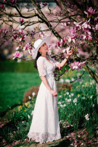 Naomie, in a white lace dress and wide-brimmed hat, stands gracefully next to a blooming magnolia tree, gently touching one of the pink flowers. Behind her is a lush green lawn adorned with delicate white blossoms.
