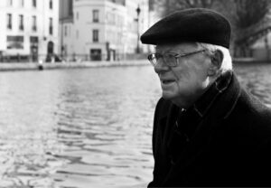 An elderly man, Vincent Levi, wearing glasses, a flat cap, and a dark coat stands by a canal with buildings in the background, looking thoughtfully into the distance. The photo is in black and white.