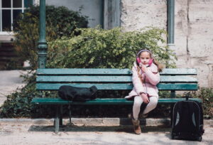 La petite Lilliane, in a pink coat and earmuffs, sits alone on a green bench outdoors. She enjoys a snack, her backpack on the ground and another bag beside her. Shrubs and an old stone wall form the backdrop.