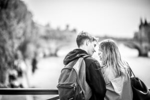 A black and white photo of a young couple with backpacks leaning close together on a bridge, lost in Le temps de l'amour, looking at each other with a blurred cityscape and river in the background.