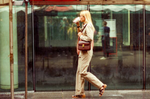 Irena Julius, a woman with long blonde hair dressed in beige and sandals, walks past a glass wall while sipping from a takeaway cup. She carries a brown shoulder bag and appears to be under a red-topped shelter.