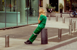 A person in green work clothes sits on a short metal post on a city sidewalk, facing away and holding their head. Glass buildings, flowers, and a blue bicycle are visible in the background.