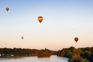 Several montgolfières float above a river surrounded by lush green trees at sunset, with a clear blue and orange sky in the background.