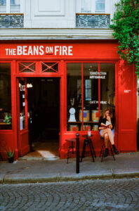 Susanna Maurice sits reading at a small table outside a vibrant red café called “The Beans on Fire” on a cobblestone street, with plants and French writing on the window.
