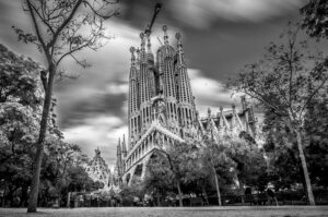 Black and white photo of the iconic Sagrada Familia basilica in Barcelone, seen from a park with trees in the foreground and dramatic clouds swirling above the church’s ornate towers.