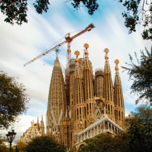 The Sagrada Familia basilica in Barcelone, Spain, features tall, ornate spires and detailed facades, with a construction crane above it and trees framing the scene under a partly cloudy sky.