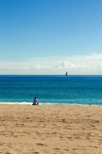 A person sits alone on a sandy beach, gazing at the blue ocean. In the distance, a sailboat glides on the water under a clear sky with a few clouds on the horizon.
