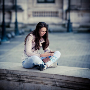 A young woman with long brown hair sits cross-legged on a stone ledge outdoors, focused on her phone. Sandra Carlois wears a light jacket and blue pants, with a notebook beside her. The blurred background hints at an urban setting.