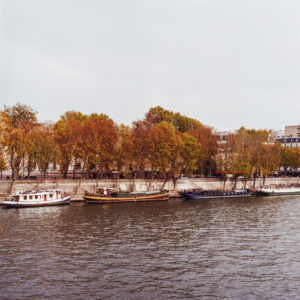 Péniches are moored along the bank of a river lined with automne-colored trees. Buildings rise behind the fiery foliage beneath a cloudy sky.