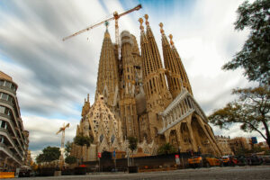 The Sagrada Familia basilica in Barcelone, Espagne, features tall spires, intricate architecture, construction cranes, and a cloudy sky in the background.