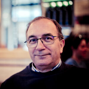 Helder VINAGRE, a middle-aged man with glasses and graying hair, smiles slightly while sitting indoors; the background is softly blurred, suggesting a casual, social setting.