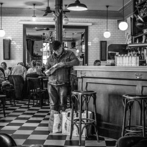 James Robert, in a denim jacket, stands near the café counter looking at his phone. Two shopping bags rest at his feet while other patrons sit at tables. The café features a checkered floor and tiled walls.