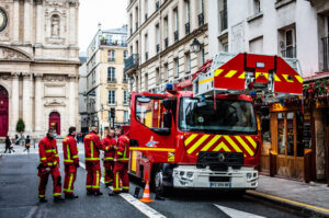 A Brigade de pompiers in red uniforms stands near a red fire truck with an extended ladder on a city street lined with buildings.