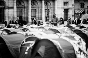 Black and white photo of people standing among rows of small tents in a city square, with a historic building featuring columns in the background. Some people wear coats and hats, suggesting cold weather.