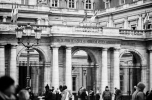 Black and white photo of people walking in front of the ornate Conseil d'État building in Paris, featuring tall arches, iron gates, and several flags above the entrance.