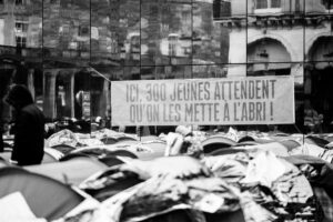 Black and white photo of tents crowding a city street. A banner on a wall reads, "ICI, 300 JEUNES ATTENDENT QU’ON LES METTE À L’ABRI !" (Here, 300 youths are waiting to be given shelter). A person stands nearby.