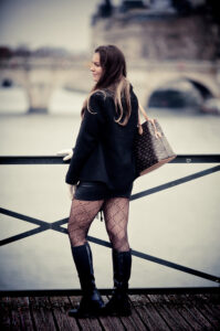 A woman with long hair stands by a riverside railing, facing away. She wears a Diane Williams black coat, patterned tights, black boots, and carries a designer bag. A blurred bridge and river are visible in the background.