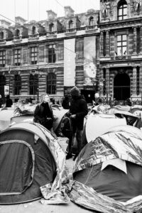 A black and white photo shows two people standing among tents covered with reflective sheets in front of a historic building, likely during a protest or demonstration. Other tents and people are visible in the background.